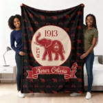 Two smiling Black women in a bedroom holding a large, custom-designed Delta Sigma Theta sorority blanket. The blanket is black with a repeating red pattern of elephants and ΔΣΘ letters, and a central circular logo featuring a large red elephant, the year 1913, stacked ΔΣΘ letters, and a red banner with the white cursive text 'Soror Olivia' and a torch.